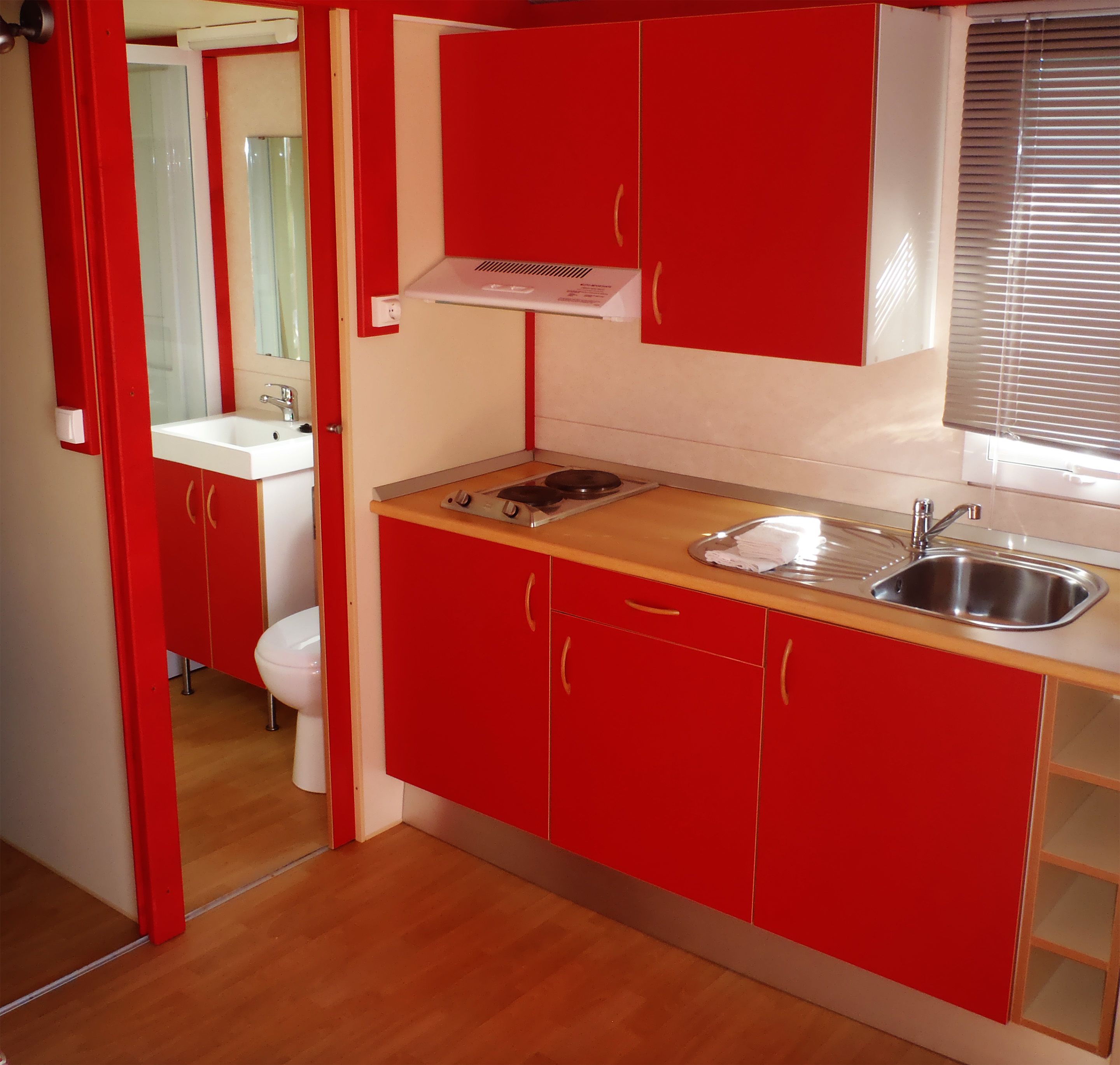 Een kleine keuken met rode kastjes, een houten aanrecht, een spoelbak en een raam met zonwering.
