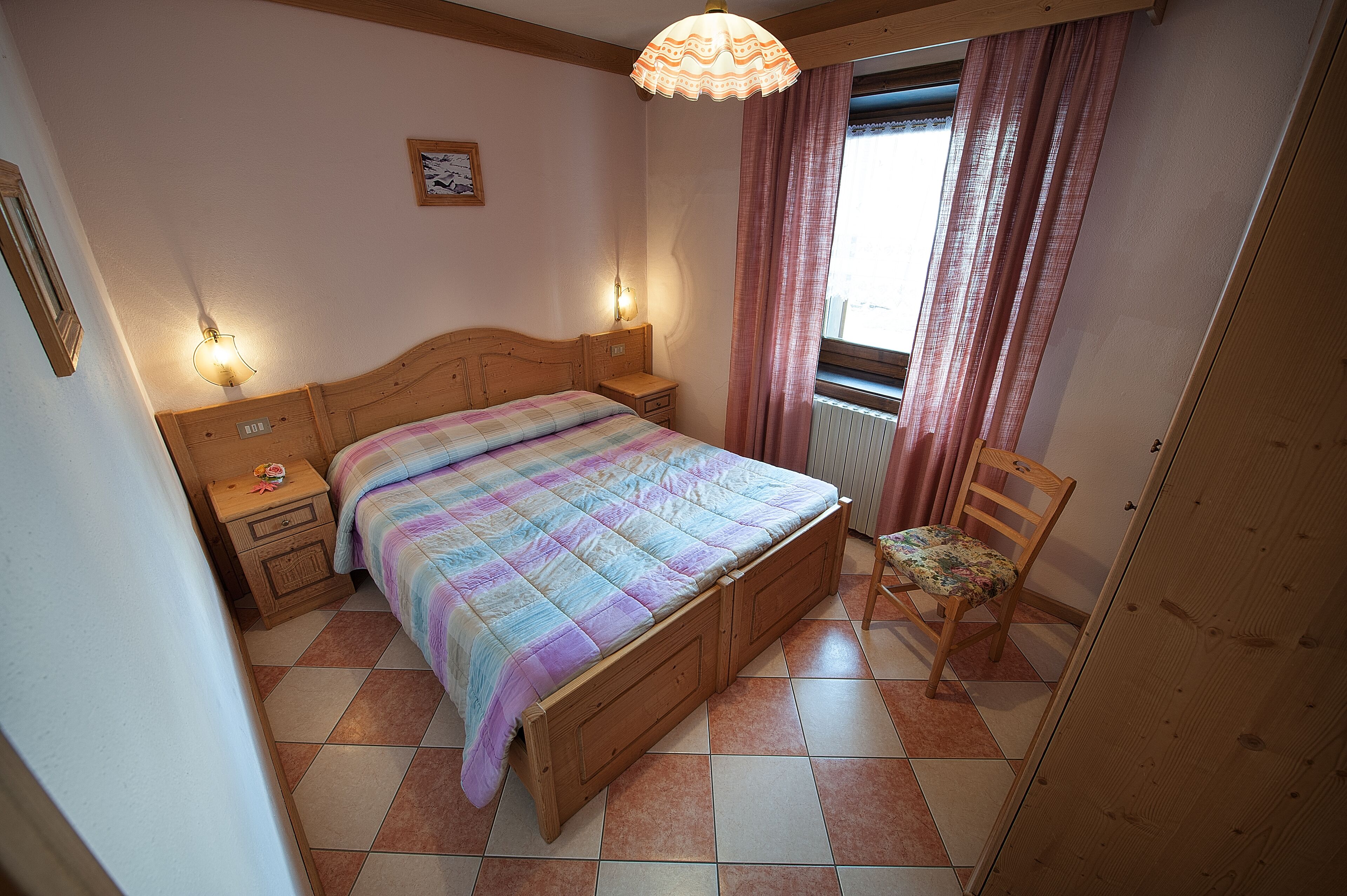 Ein Schlafzimmer mit einem hölzernen Bett, zwei Nachttischen, einem Beistelltisch, einem Stuhl, einem Fenster mit Vorhängen und einer Pendelleuchte.