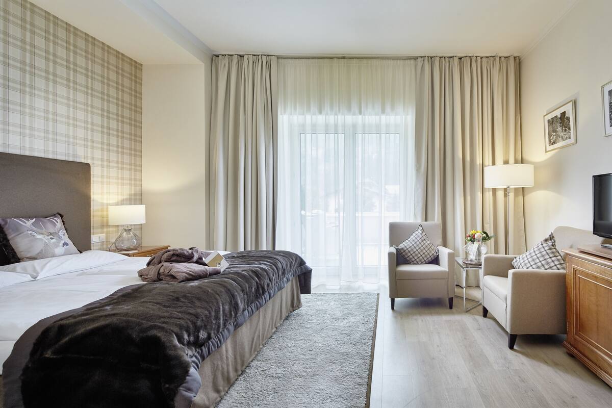 Una habitación de hotel con cama, sofá, televisor, lámpara y ventana con cortinas.