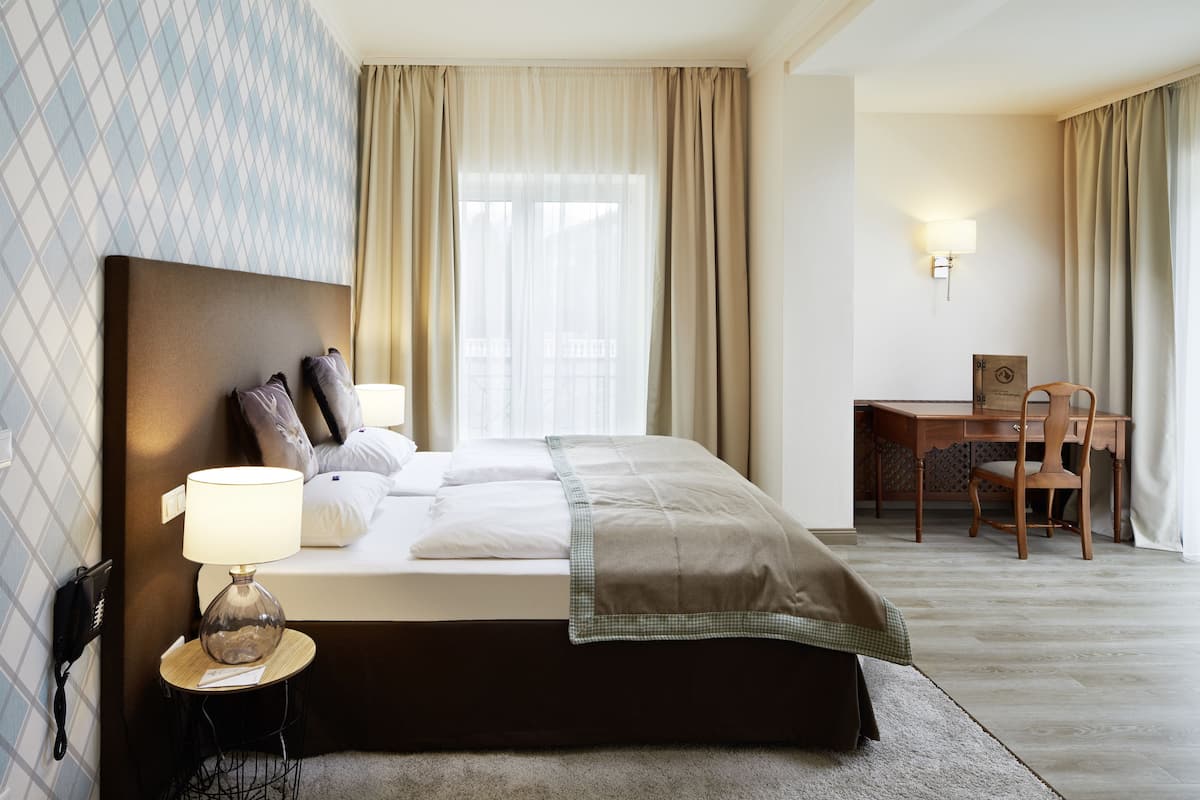 Habitación de hotel con cama, lámpara de noche, escritorio, silla y ventana con cortinas.
