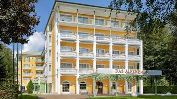 Un hotel de varios pisos con fachada amarilla, múltiples balcones y un letrero que dice "Das Alpenhaus".