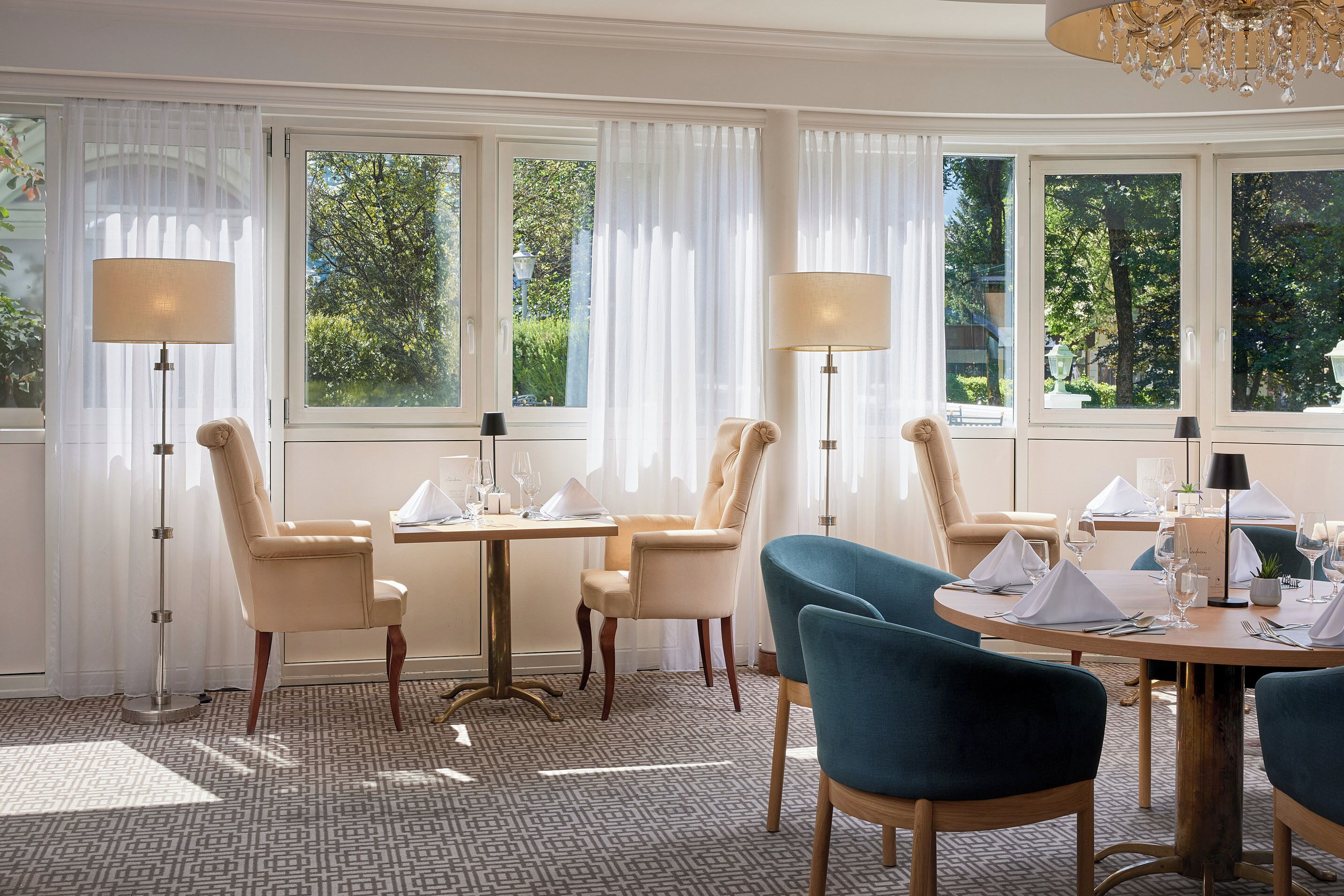 A dining area with a round table set for four, surrounded by chairs, large windows with sheer curtains, and floor lamps.