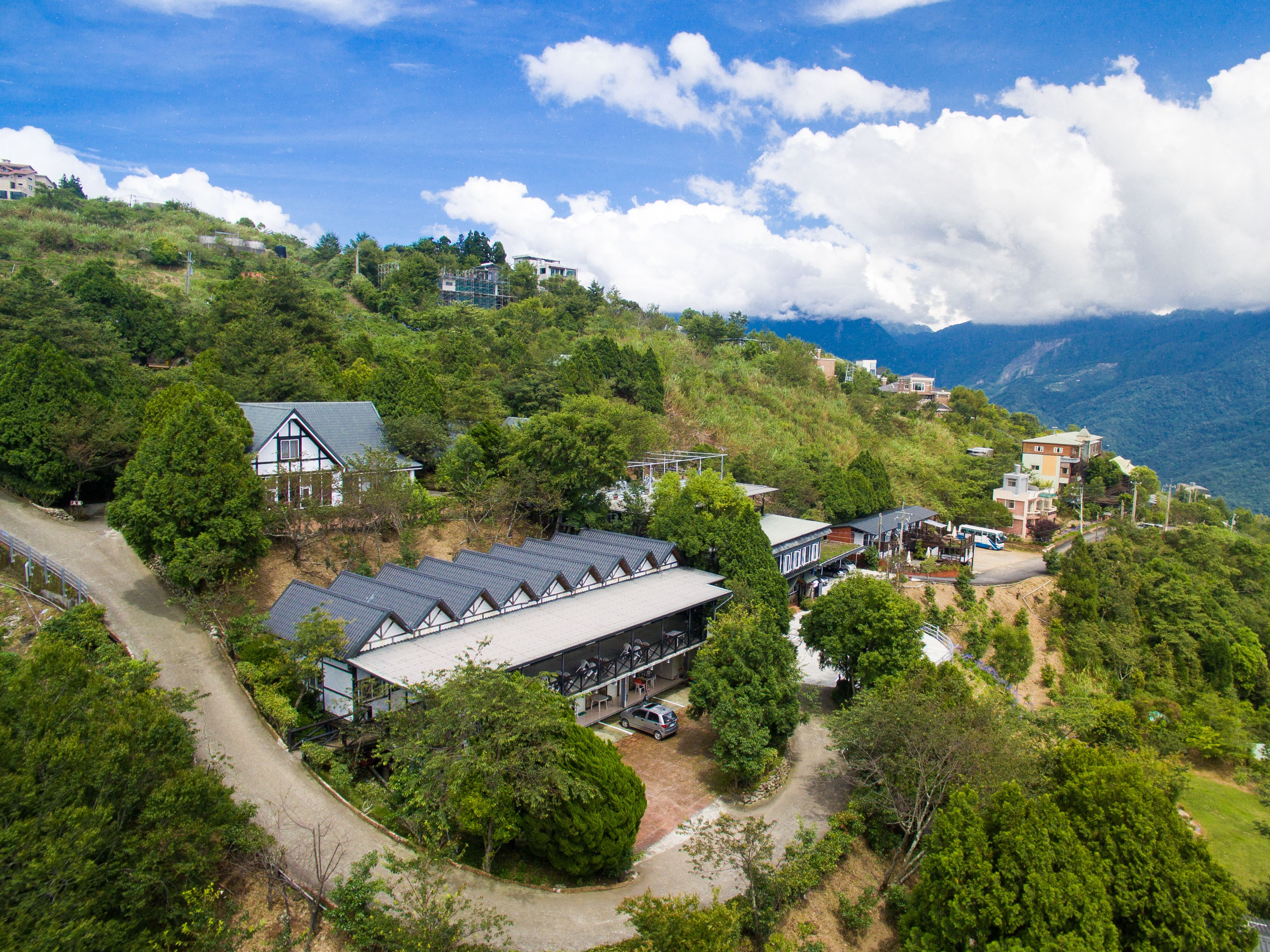 A hillside property with multiple buildings, including a large structure with a sloped roof, surrounded by lush greenery and a clear sky.