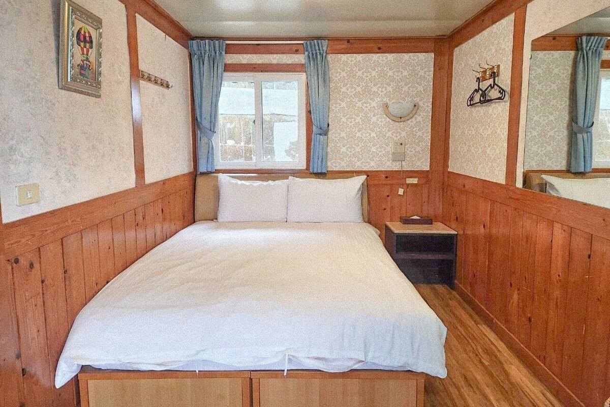 A wooden-paneled room with a bed, a nightstand, and a window with curtains.