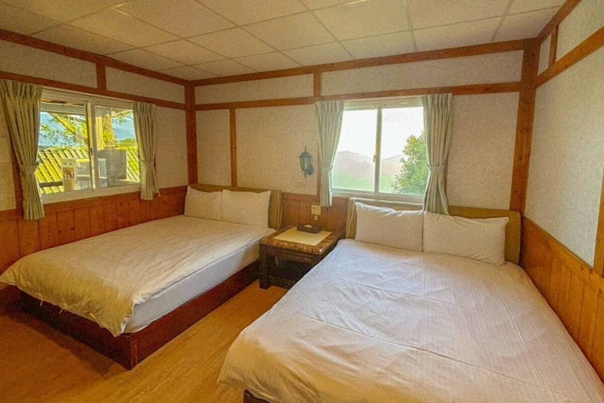 A room with two single beds, wooden walls, a small table, and windows with curtains.