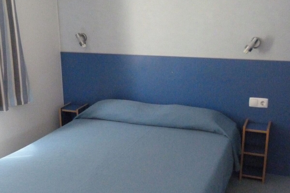 Una habitación con cama, una mesita, una silla, un enchufe y una ventana con cortinas.