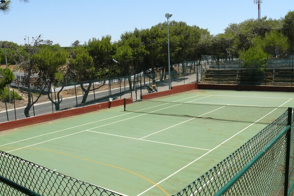 Una cancha de tenis al aire libre con red, rodeada por una cerca de malla y árboles.