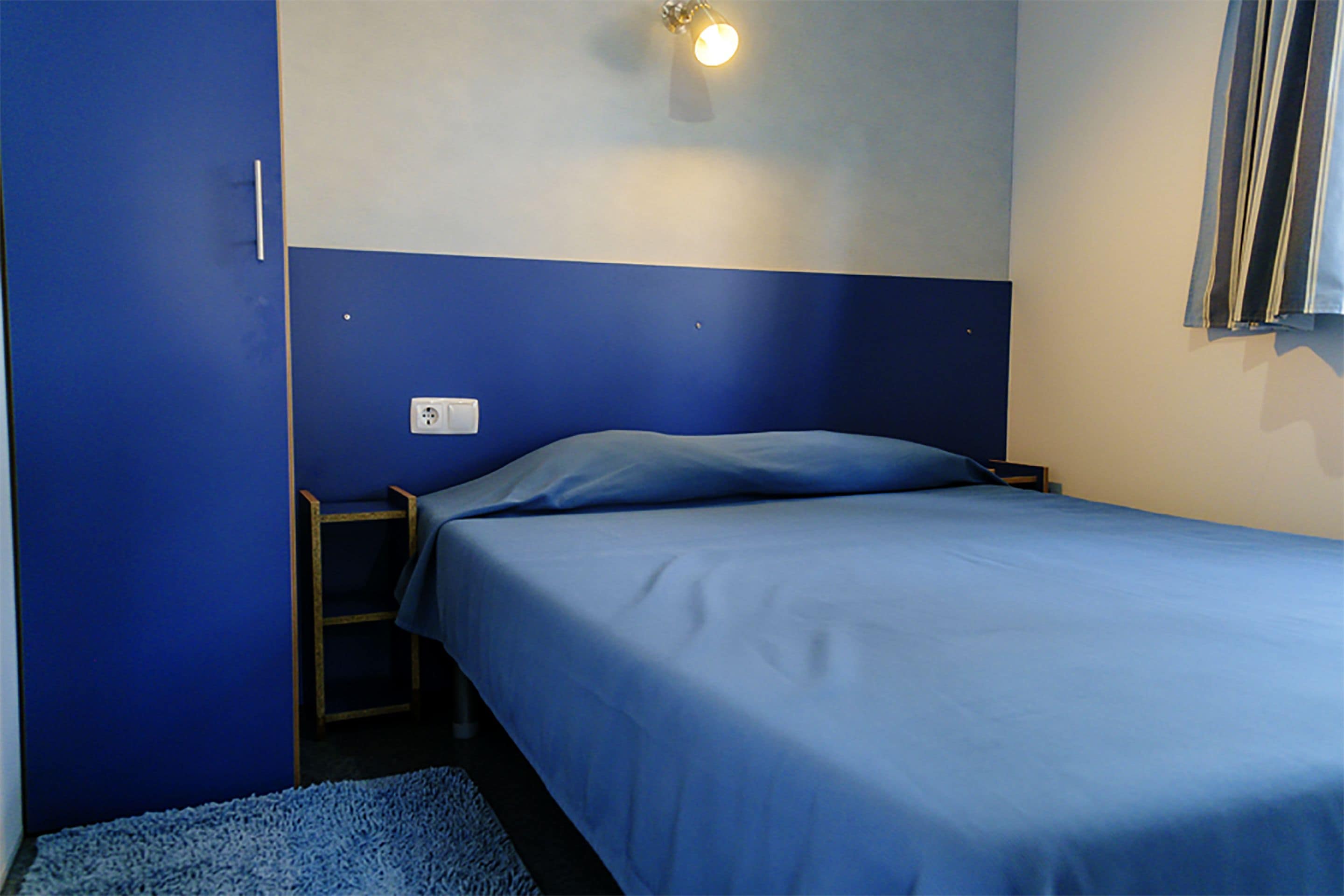 Una cama individual con colcha y almohada azules en una habitación con paredes azules y techo blanco. Junto a la cama, una mesita de noche de madera con una lámpara.