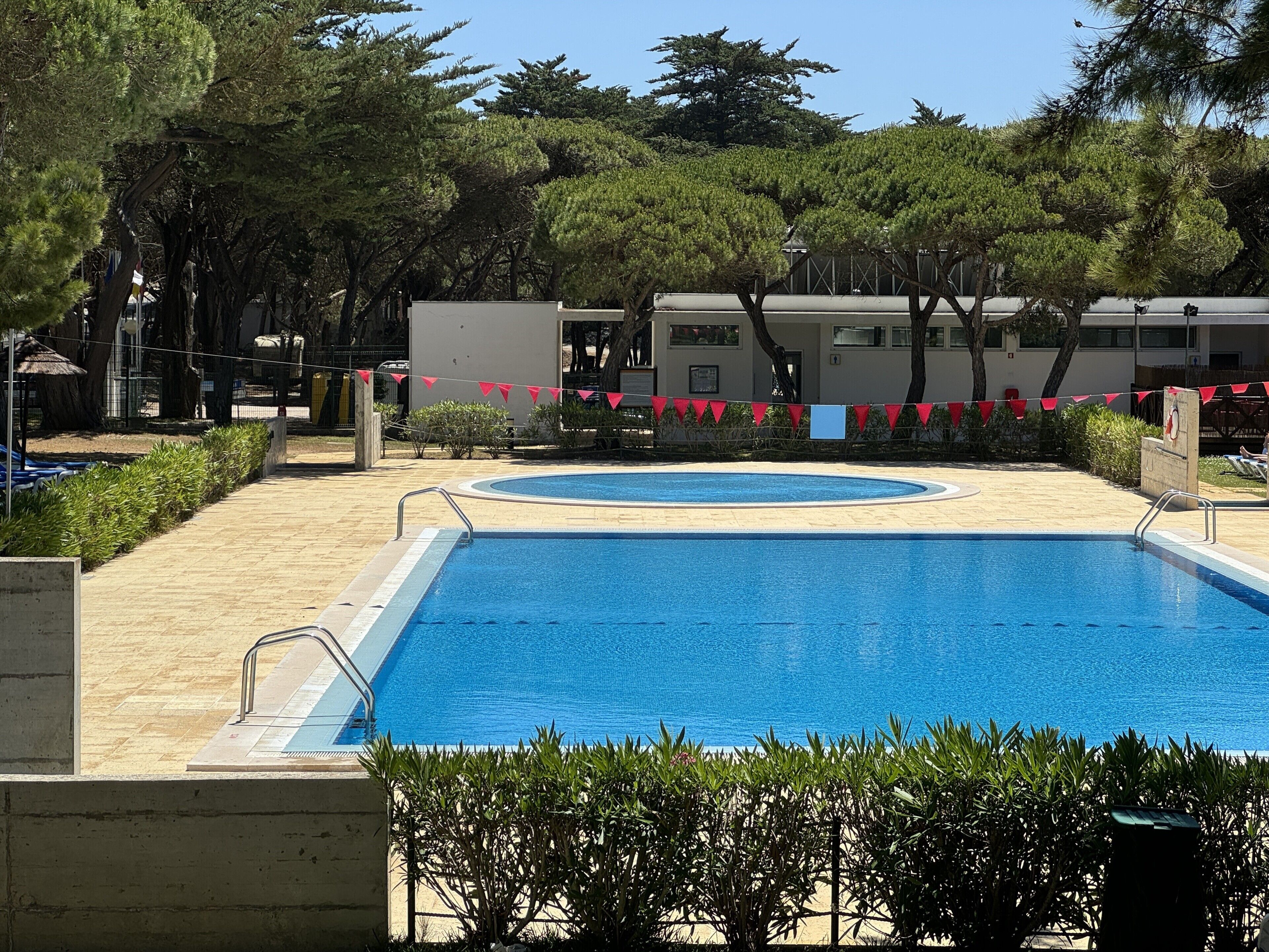 A rectangular swimming pool with a surrounding tiled deck, surrounded by trees and a building with a balcony.