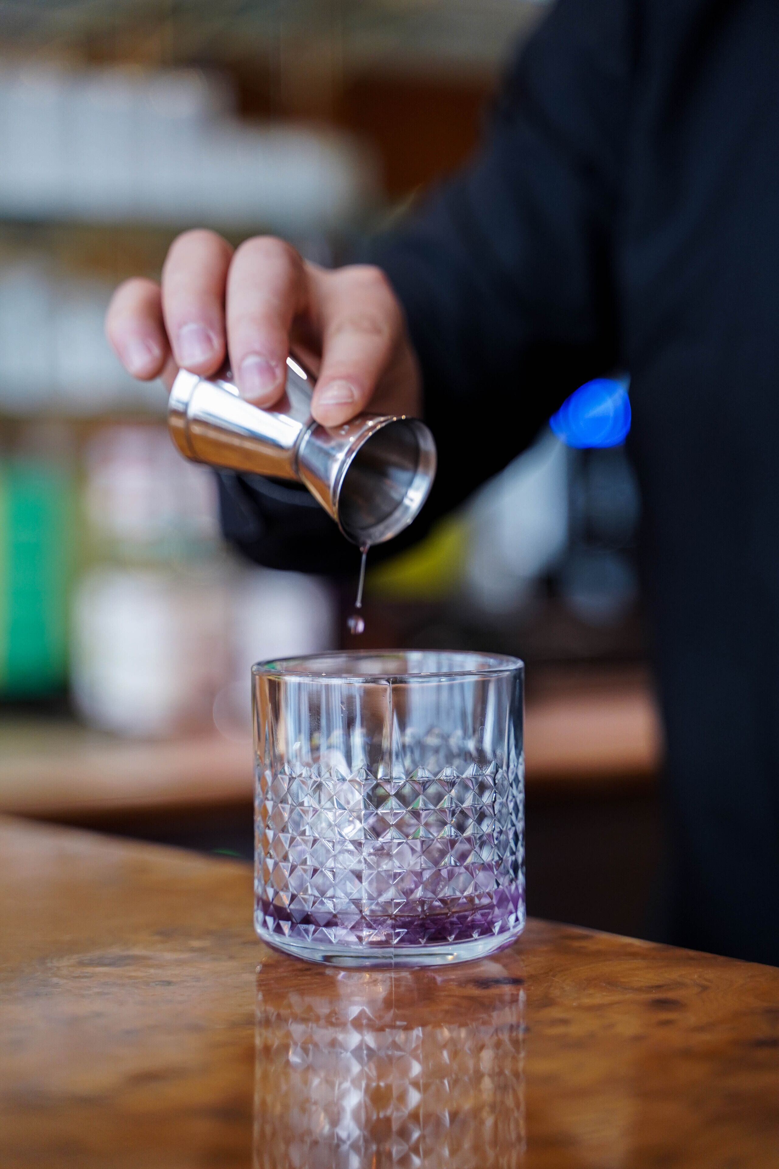 A hand pouring a liquid from a metal jigger into a clear glass with a textured pattern.