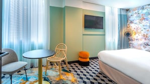 A hotel room with a bed, a round table, a chair, a small orange ottoman, and a TV on the wall.