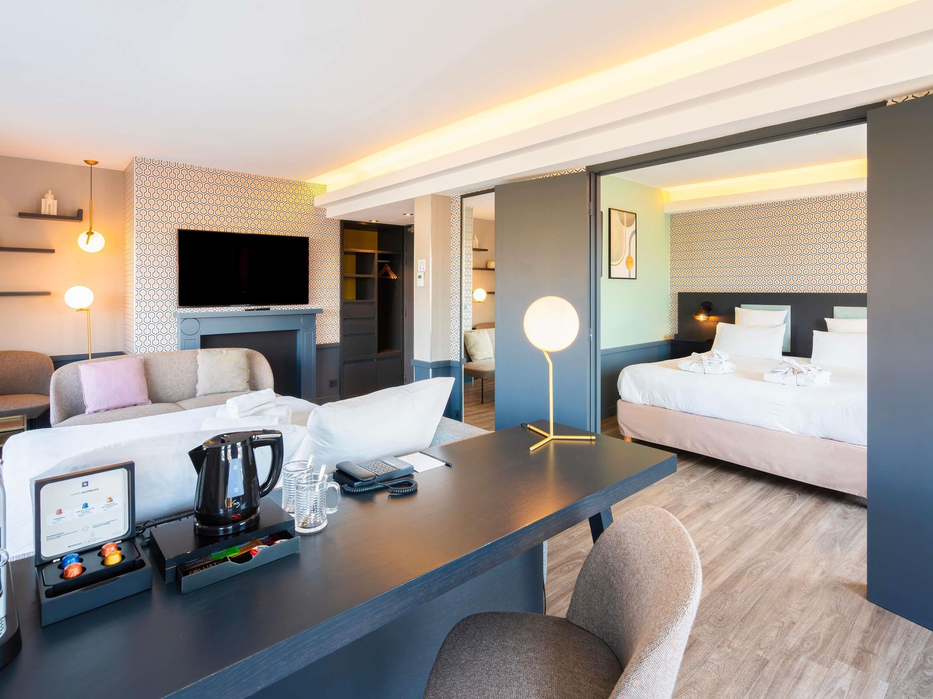 A modern hotel room with a sofa, a desk with a coffee maker, a flat-screen TV, and a bed.