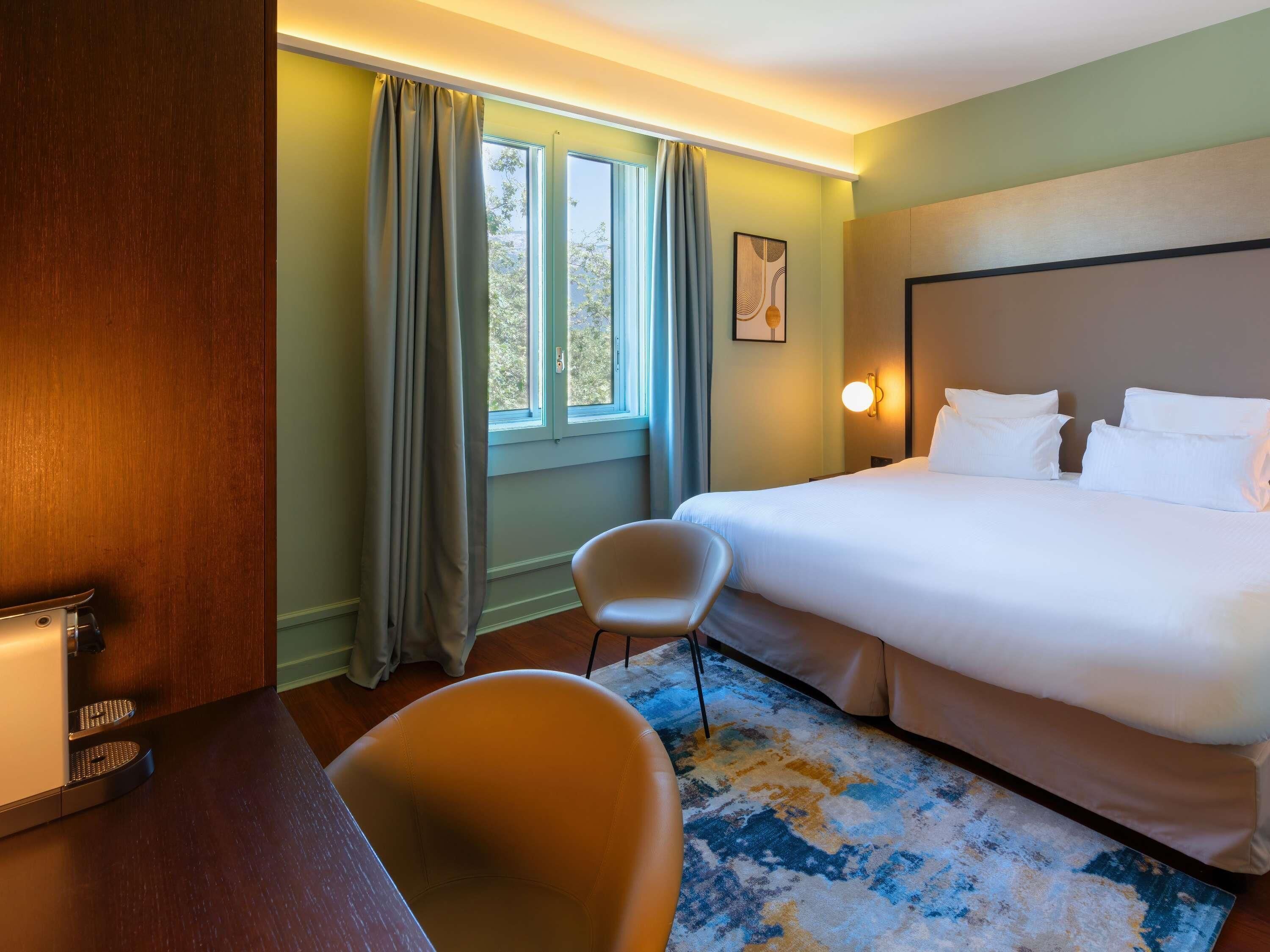 A hotel room with a bed, a chair, a desk, a window with curtains, and a wall-mounted light.