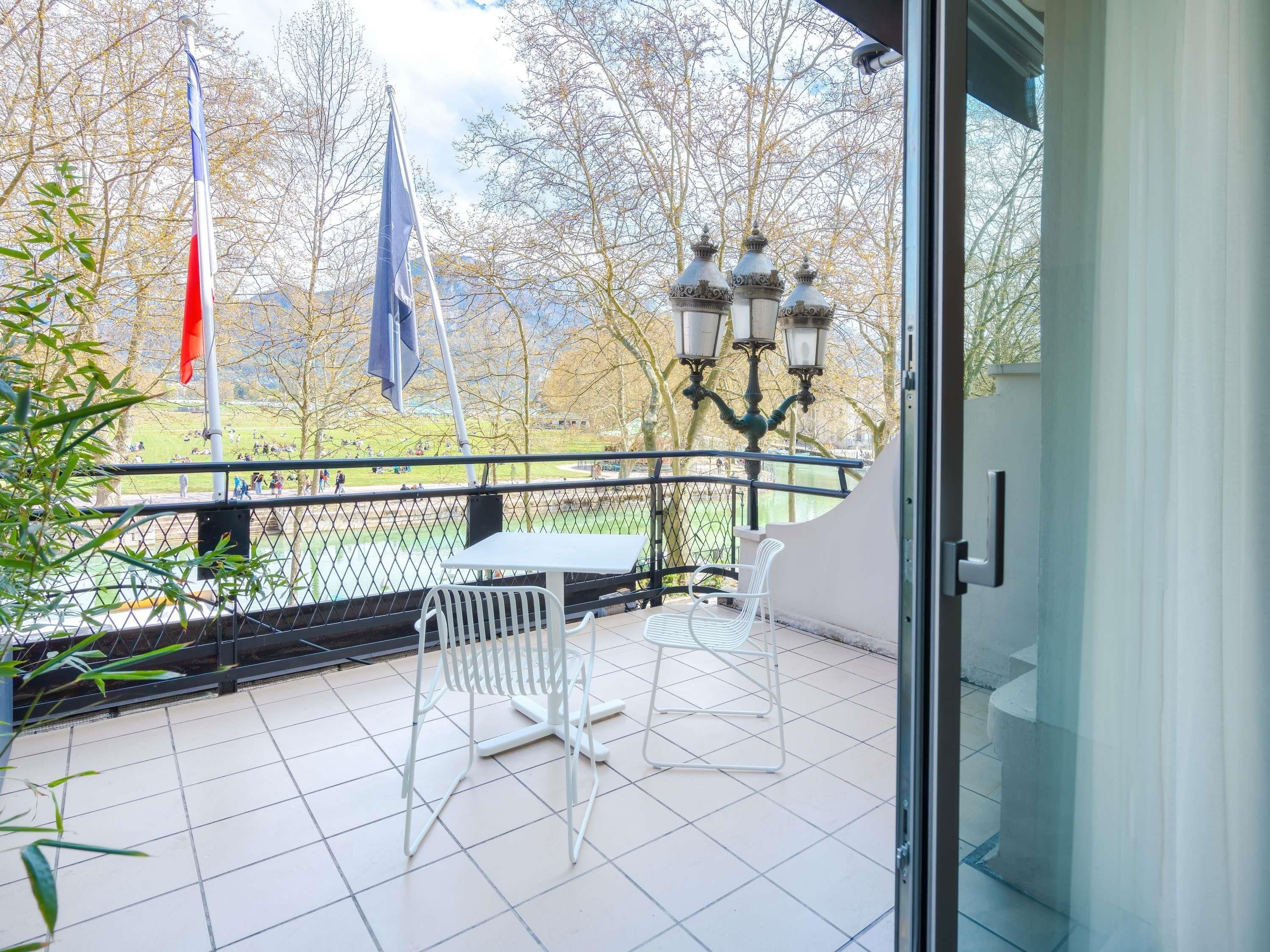A balcony with a table and chairs, a view of a park with trees and a body of water, and flags.