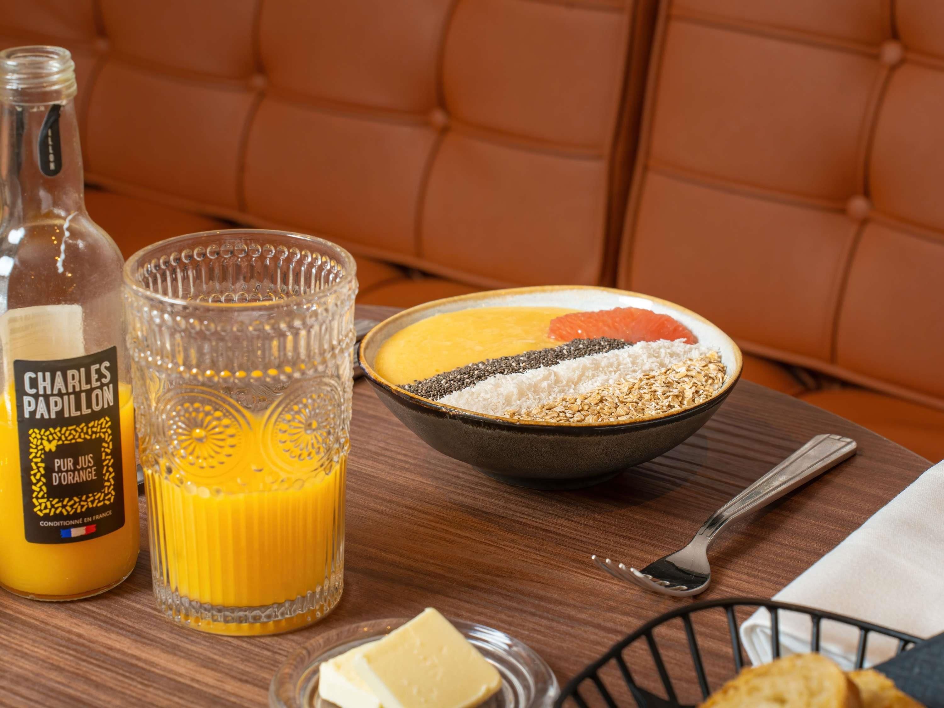 A breakfast setting with a bottle of orange juice, a glass, a bowl of cereal with fruits and seeds, a butter dish, and a bread basket.