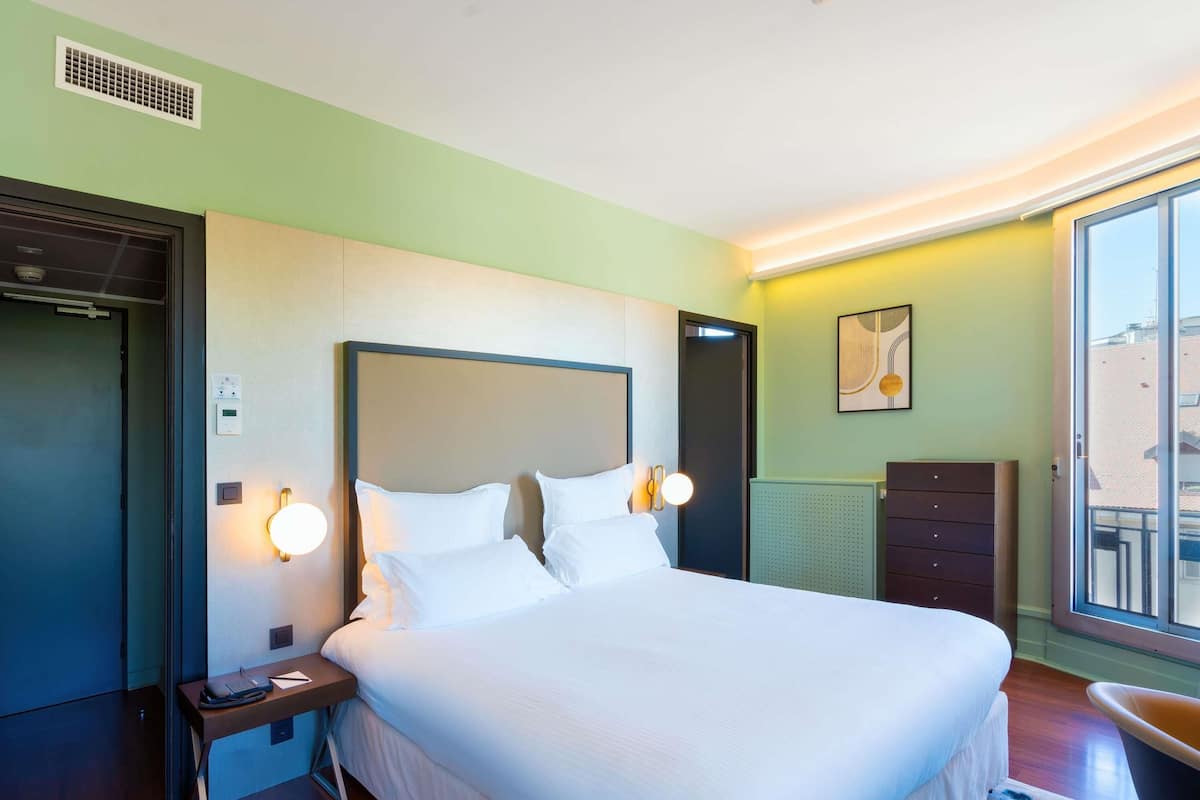 A hotel room with a large bed, bedside lamps, a desk with a telephone, and a view of the outside through a window.