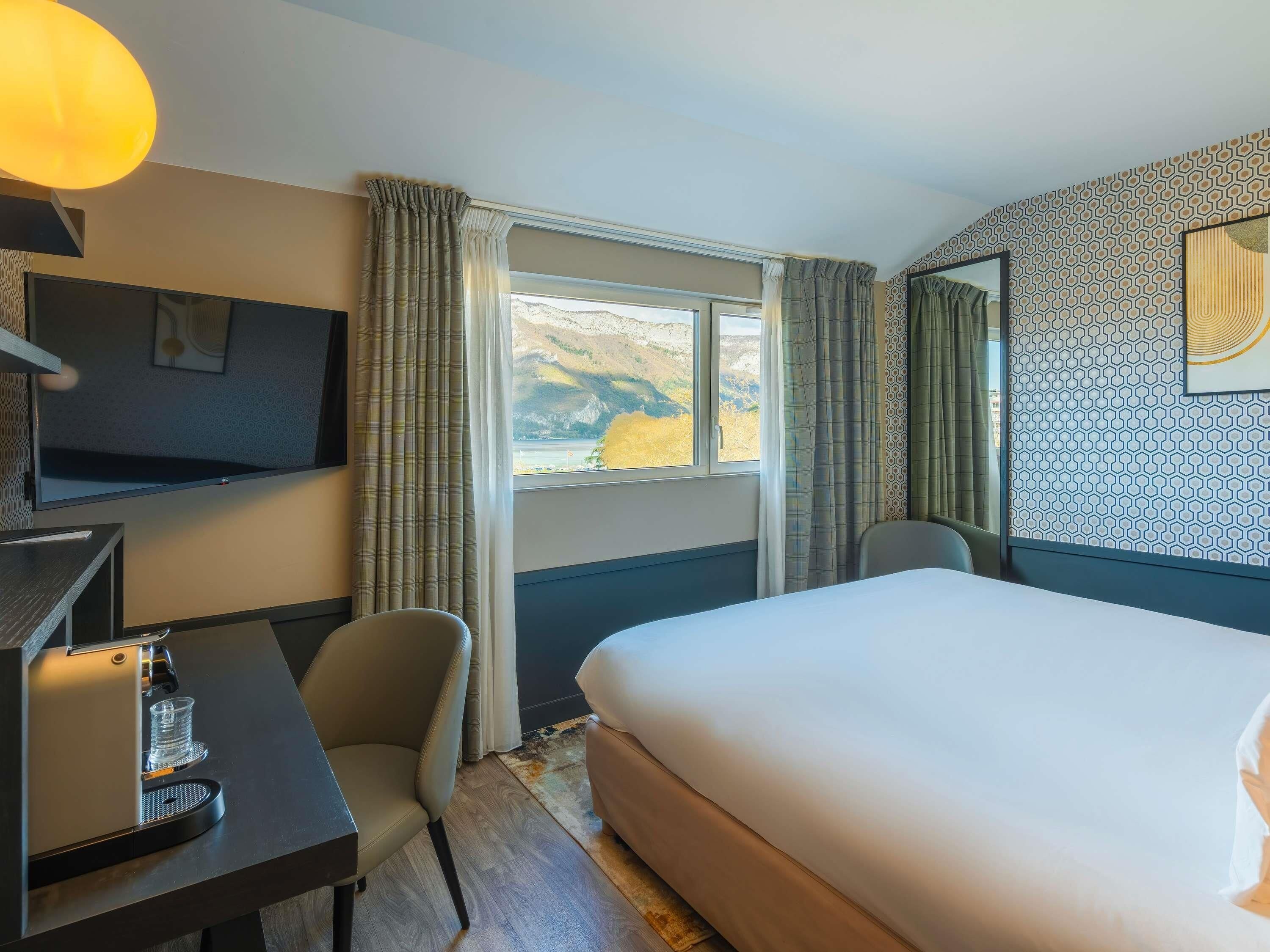 A hotel room with a bed, a desk with a coffee machine, a TV, and a window with curtains.