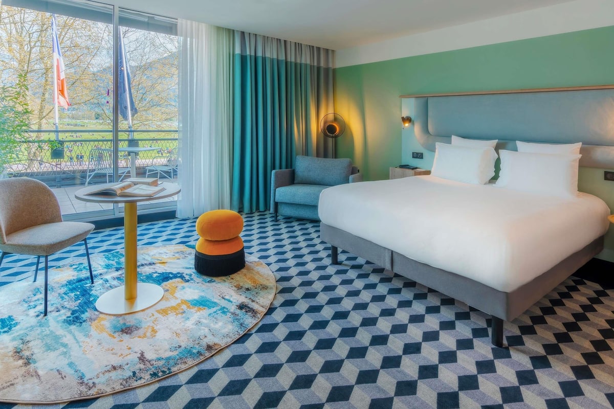 A hotel room with a bed, a chair, a small table, and a view of the outdoors.