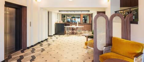 Un hall de hotel con piso estampado, un sillón amarillo, una mesa redonda con una revista y una zona de bar al fondo.
