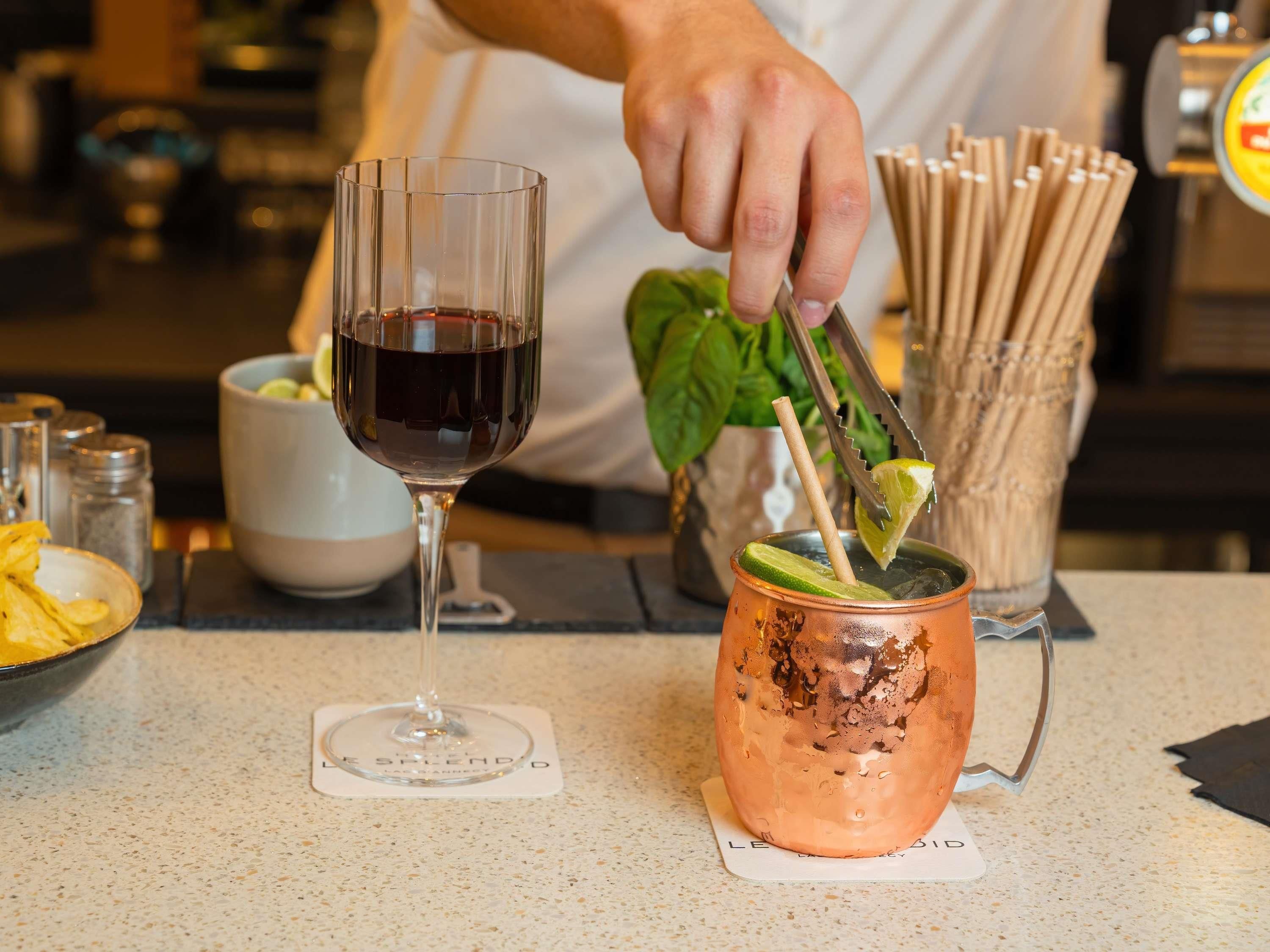 A person preparing a drink with a lime slice and a straw in a copper mug on a table.