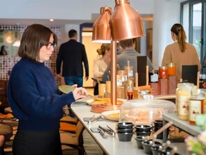 A person serving food at a counter with a copper lamp, jars, and utensils.