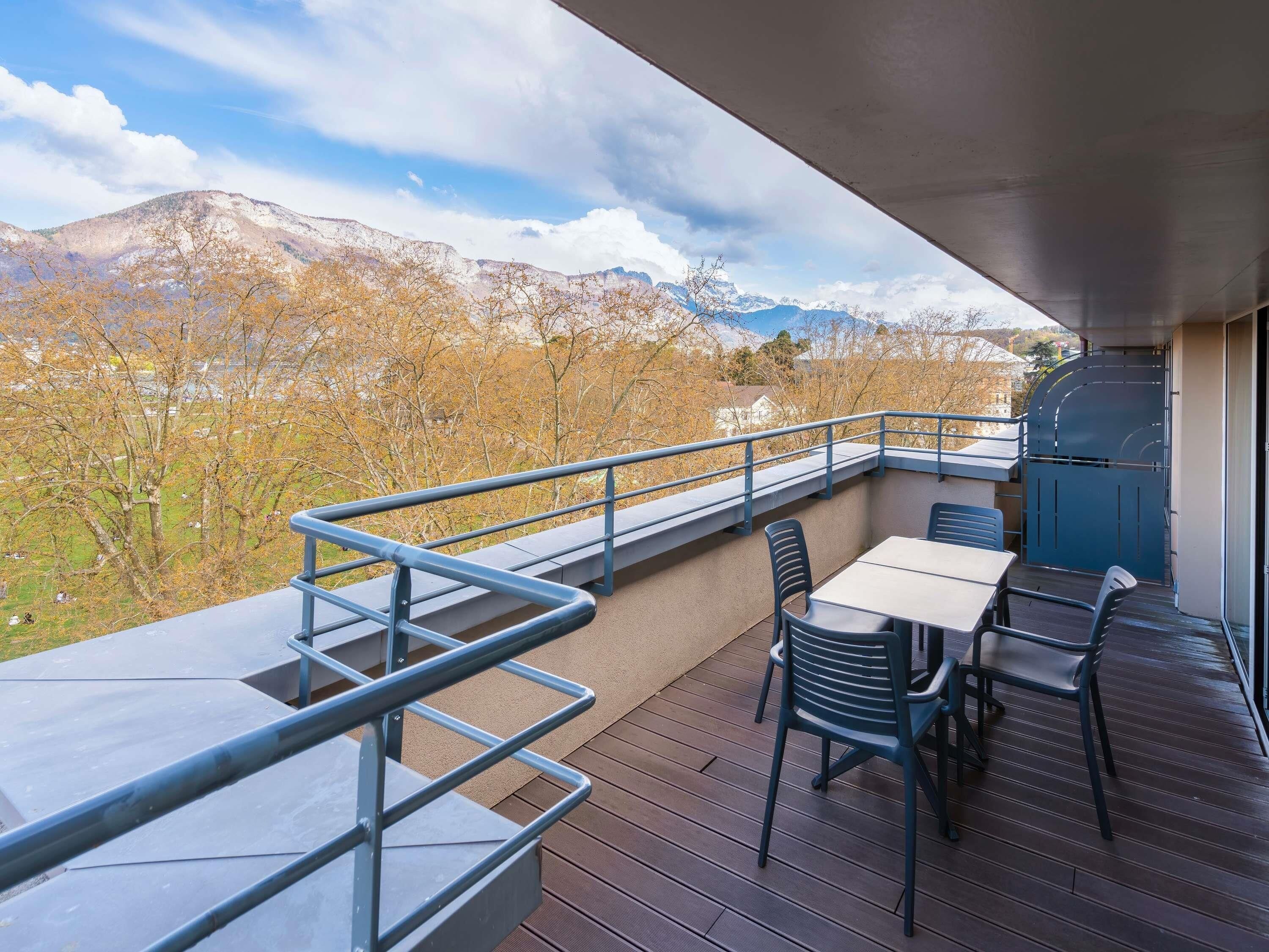 An outdoor balcony with a table and chairs, a metal railing, and a view of mountains.