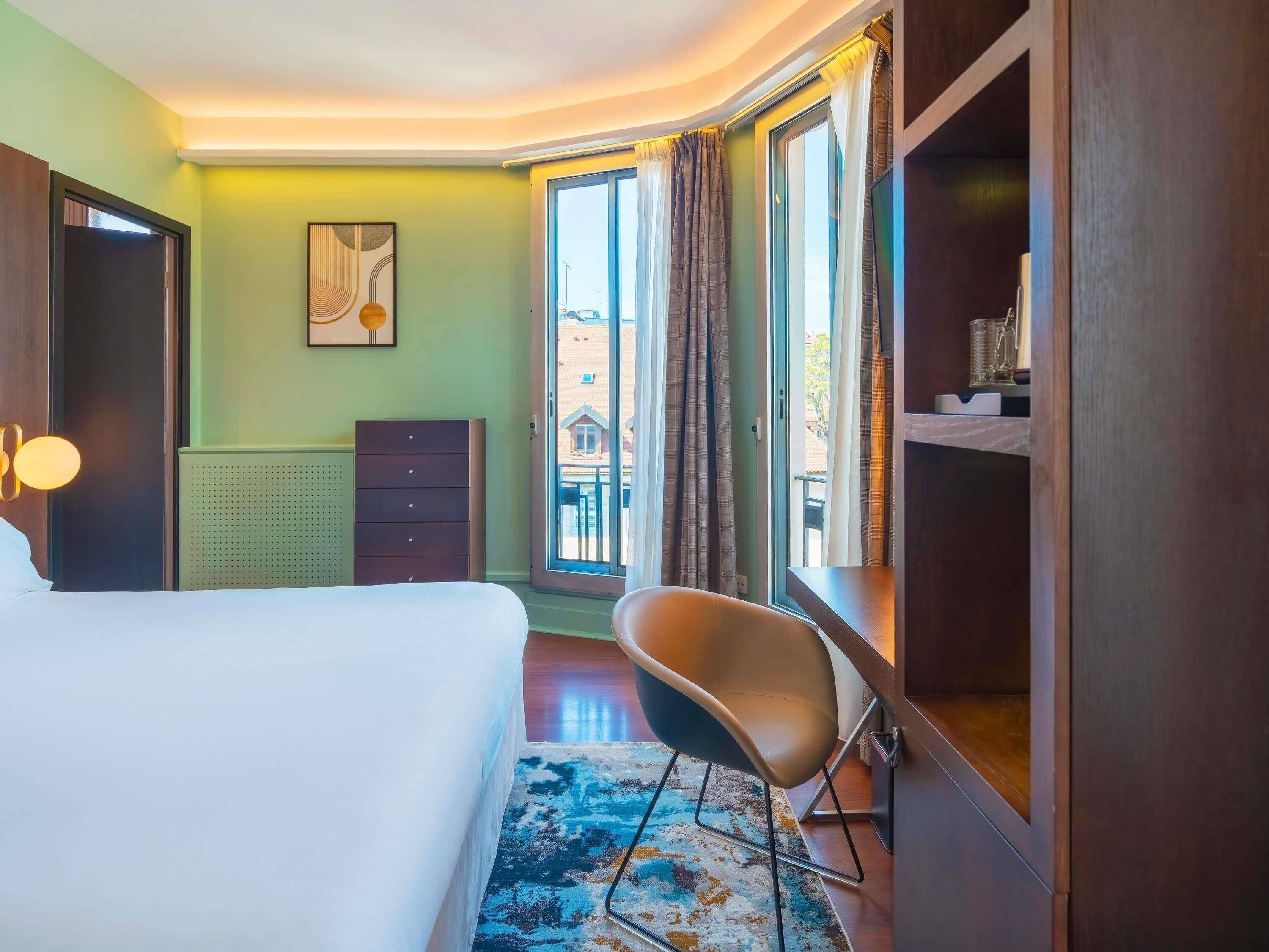 A hotel room with a bed, a chair, a dresser, a radiator, a window with curtains, and a view of buildings outside.