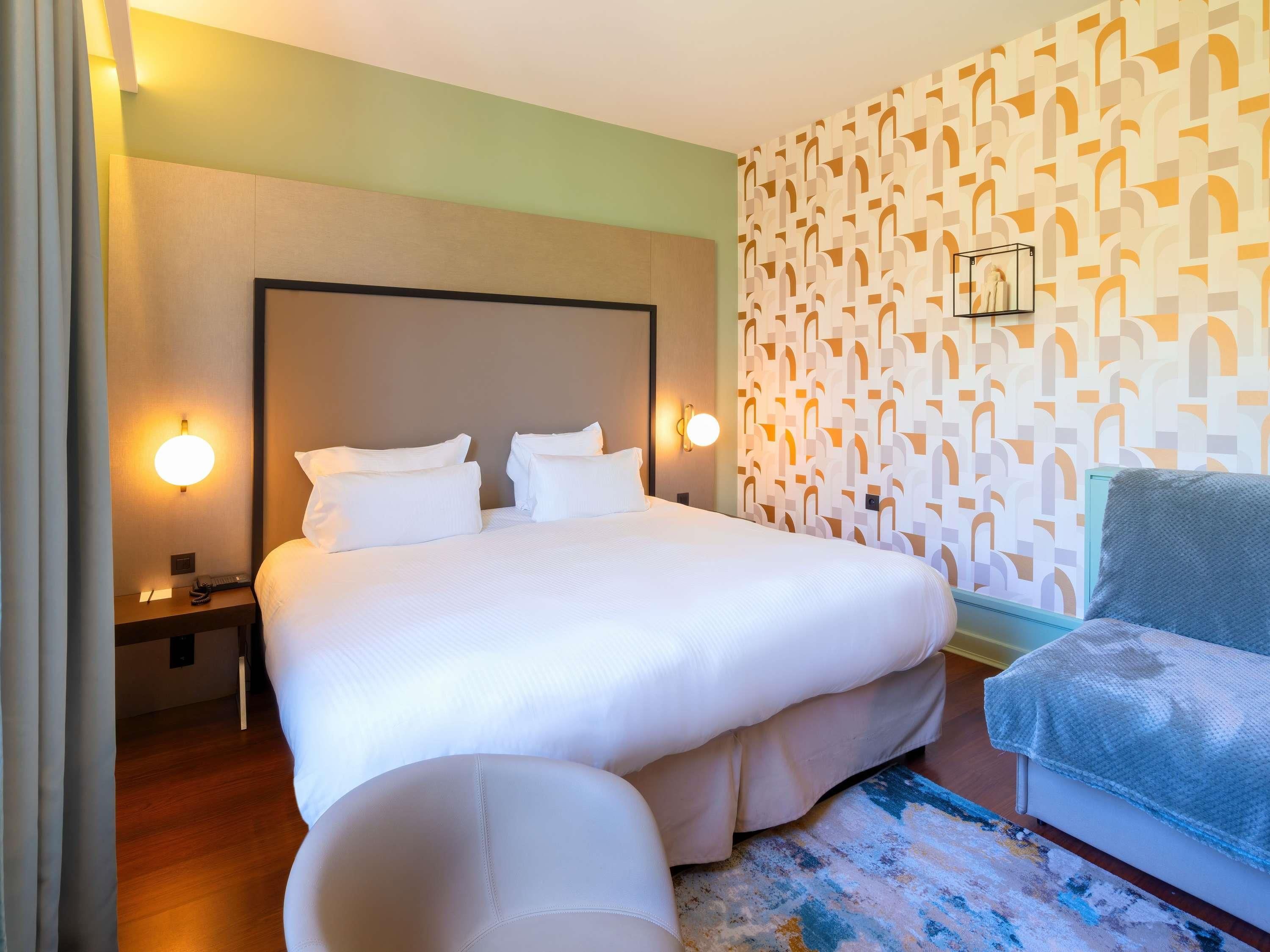 Una habitación de hotel con una cama grande, lámparas de noche, un papel tapiz estampado y un sillón azul.