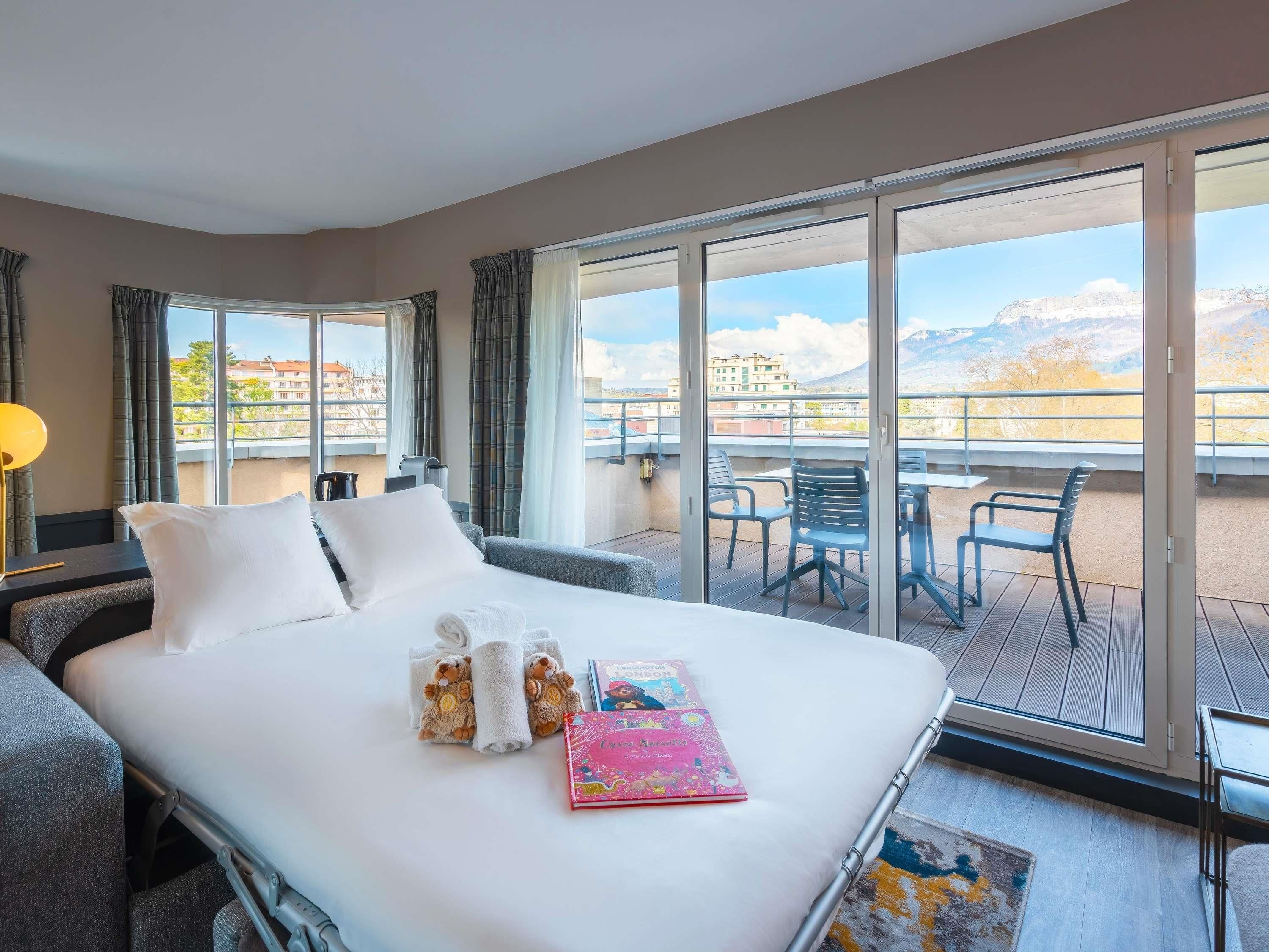 Habitación de hotel con una cama, un balcón con sillas y vistas a edificios y montañas.