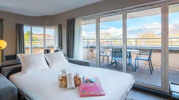 Habitación de hotel con una cama, un balcón con sillas y vistas a edificios y montañas.