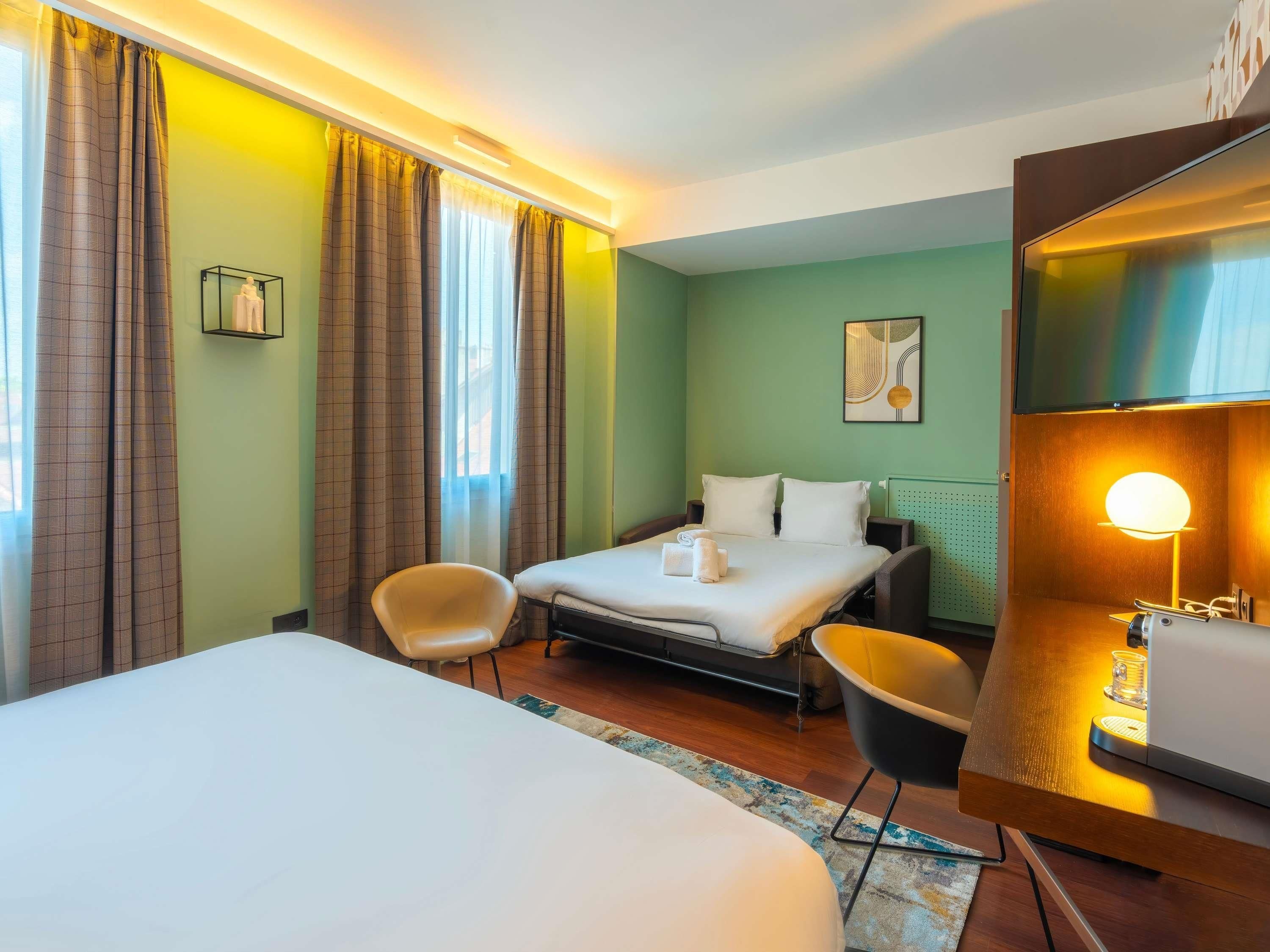 Una camera d'albergo con un letto, due poltrone, una scrivania con una lampada e una finestra con tende.
