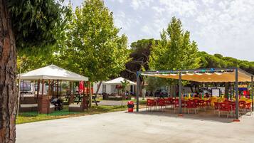 An outdoor seating area with tables and chairs, a covered picnic area, and a tree in the background.