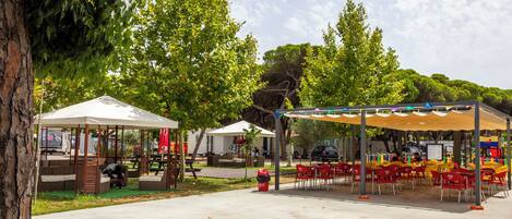 An outdoor seating area with tables and chairs, a covered picnic area, and a tree in the background.