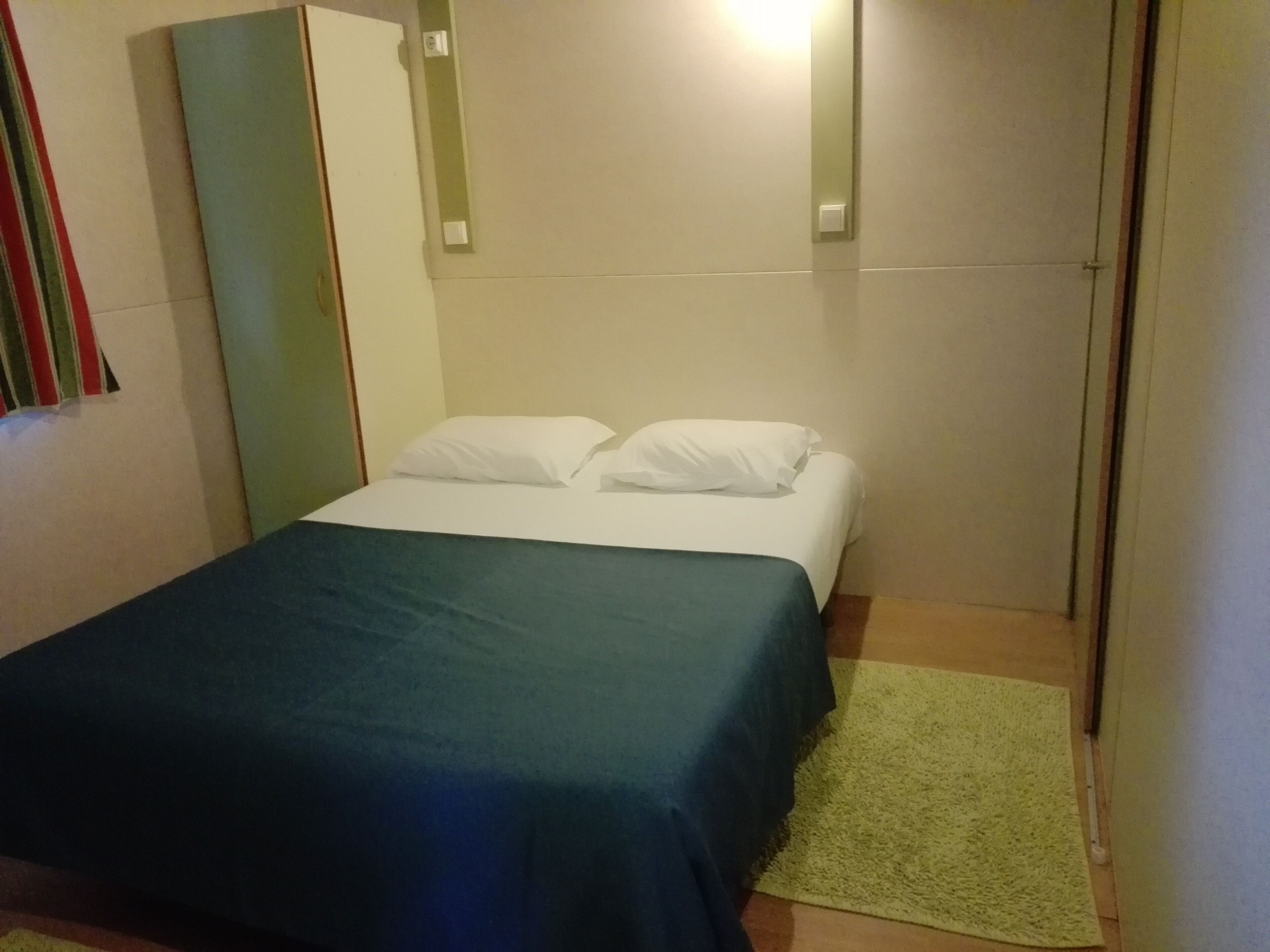 A small, single-bed room with a green bedspread, white pillows, and a green rug.