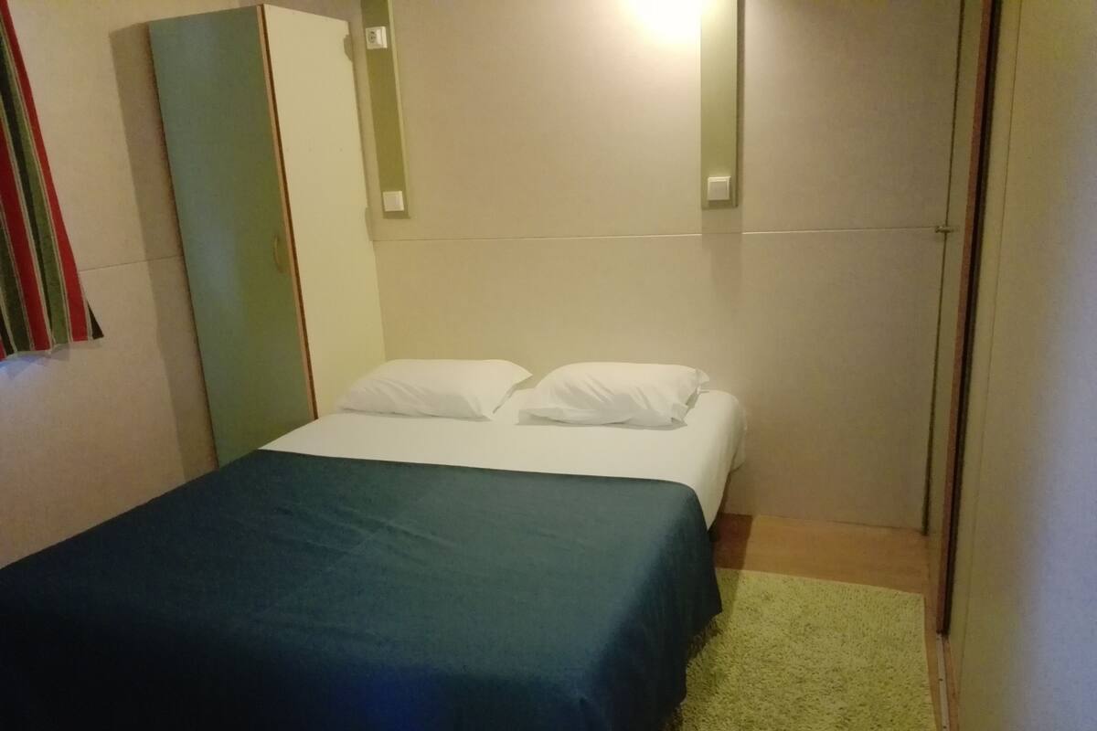 Habitación pequeña con una cama individual, colcha verde, almohadas blancas y alfombra verde.