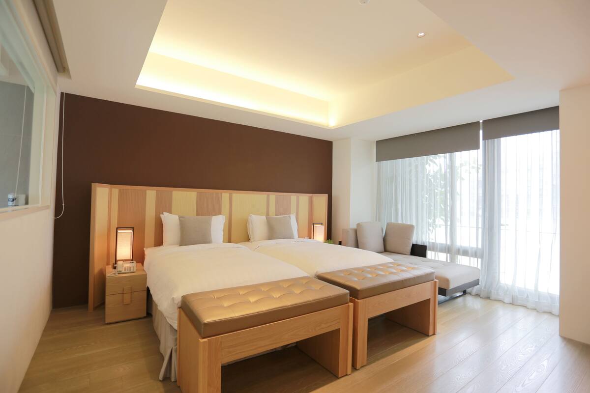 Una habitación de hotel moderna con una cama grande, mesitas de noche, un banco y un ventanal con cortinas.