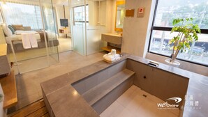 Une chambre d’hôtel moderne dotée d’une paroi en verre séparant la chambre à coucher de la salle de bain, d’une grande fenêtre offrant une vue dégagée et d’un petit coin salon agrémenté d’une plante.