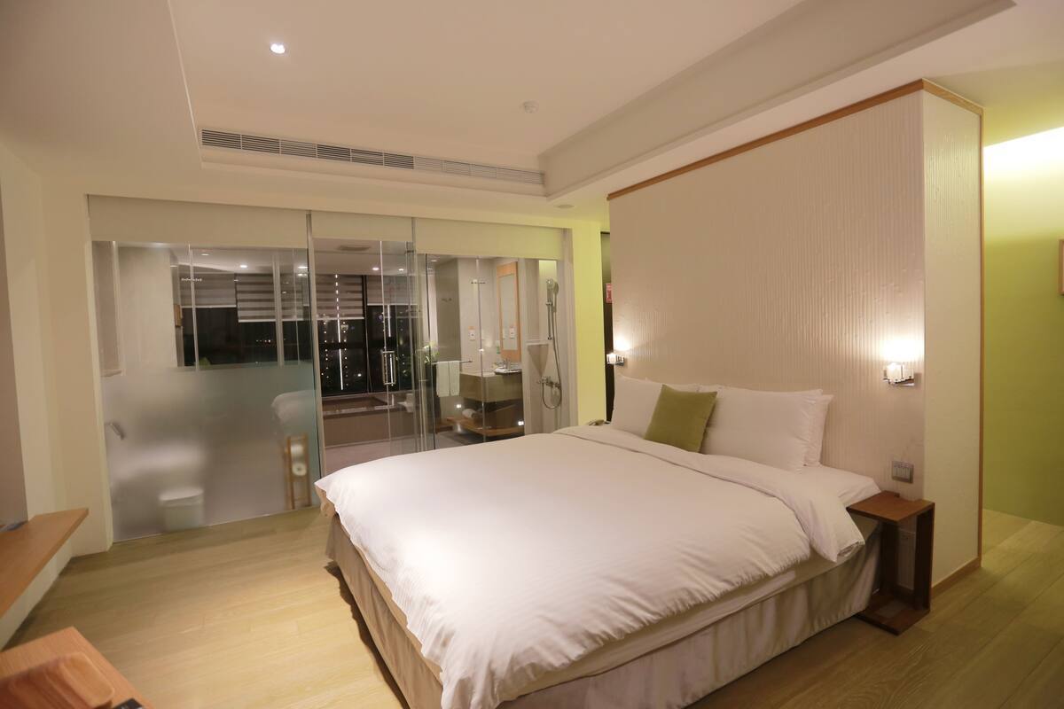 Habitación de hotel moderna con una cama grande, cabina de ducha de vidrio y vistas a la ciudad de noche.