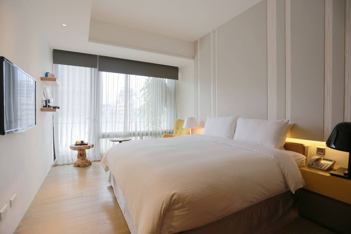 Habitación de hotel con una cama grande, mesita de noche con lámpara, una pequeña mesa redonda con espejo y frutero, y una ventana con cortinas translúcidas.