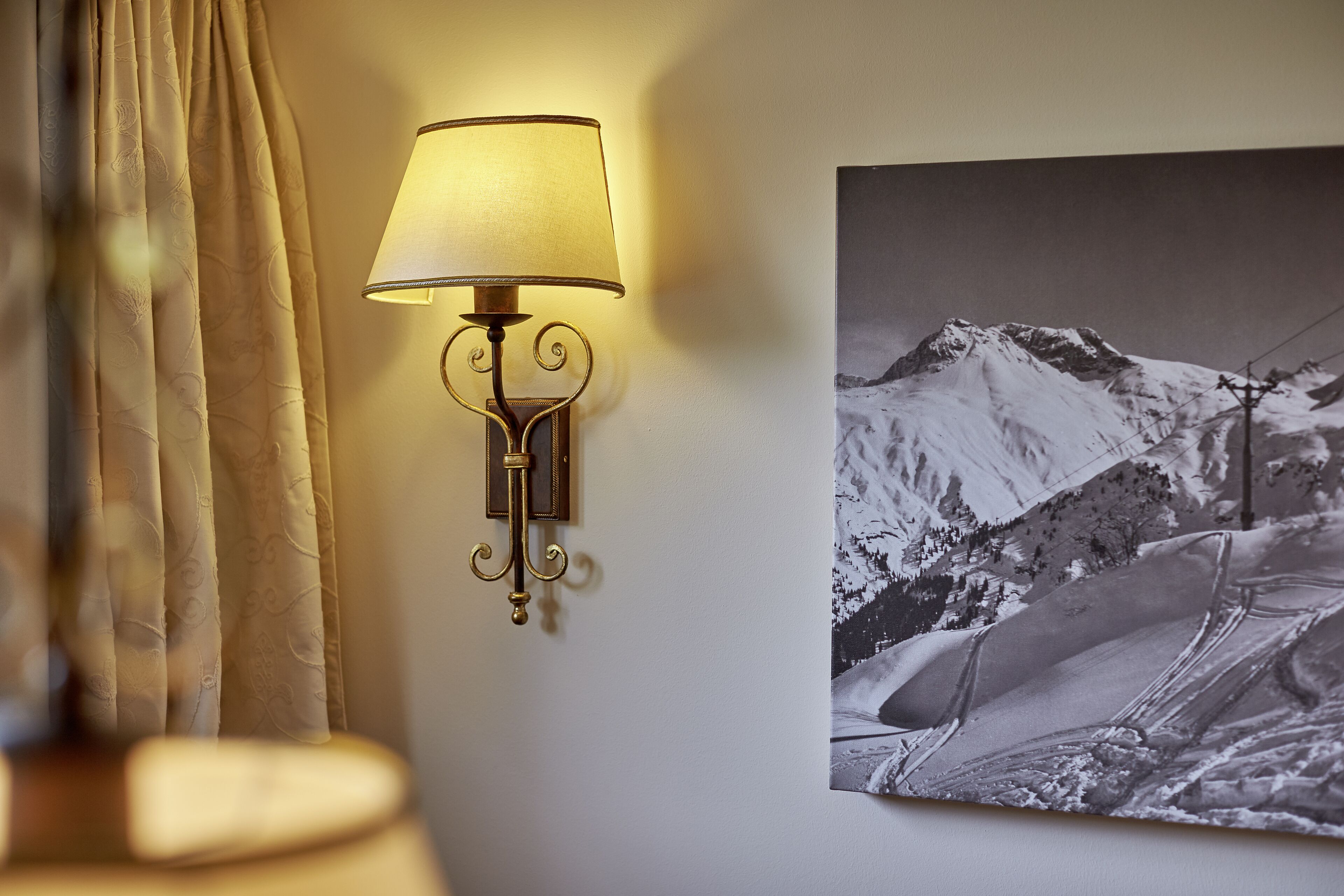 A wall-mounted lamp with a decorative design, a framed photograph of a snowy mountain landscape, and a curtain partially visible.