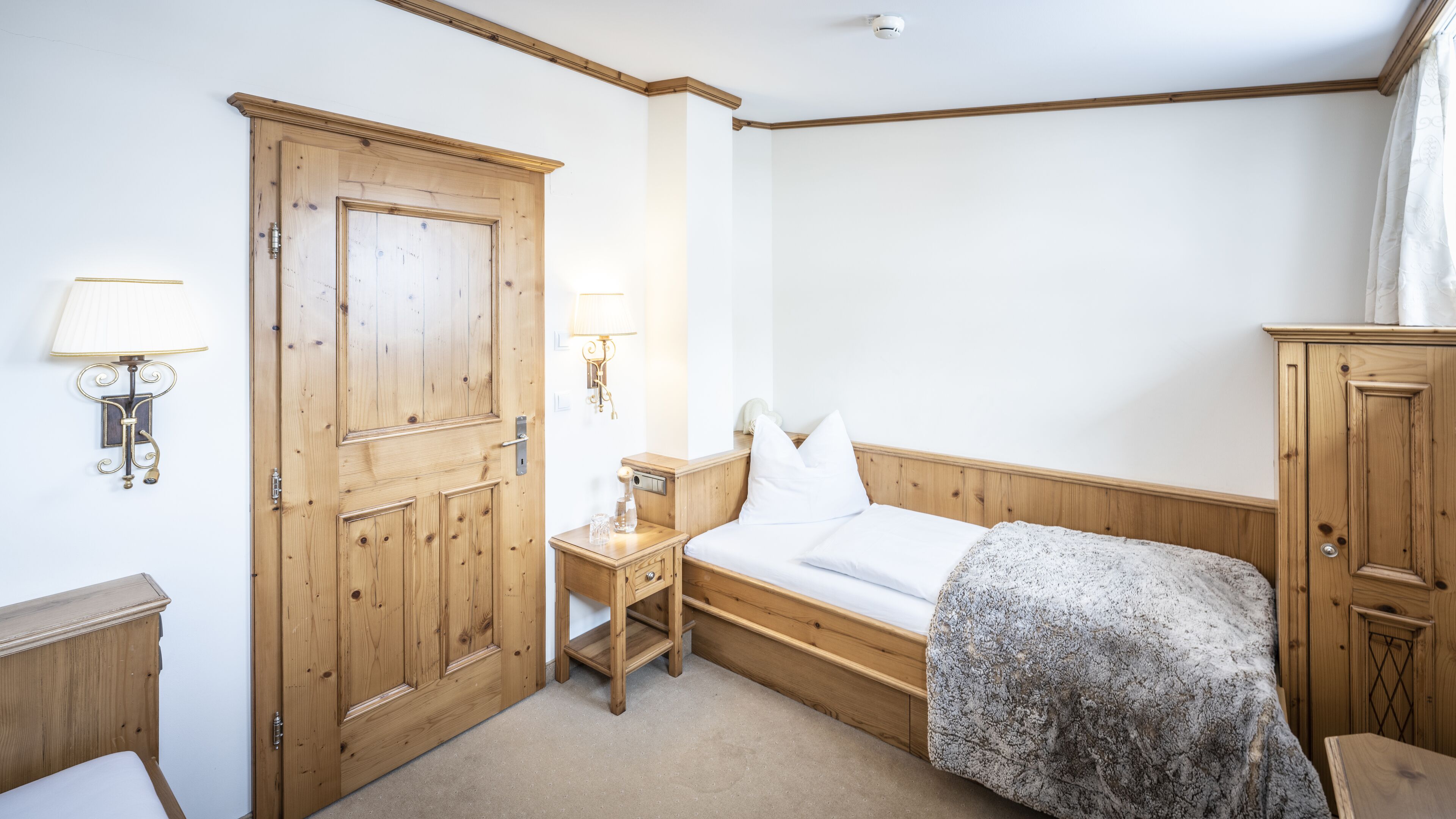 A room with a wooden door, a bedside table with a lamp, a bed with a blanket, and a wardrobe.