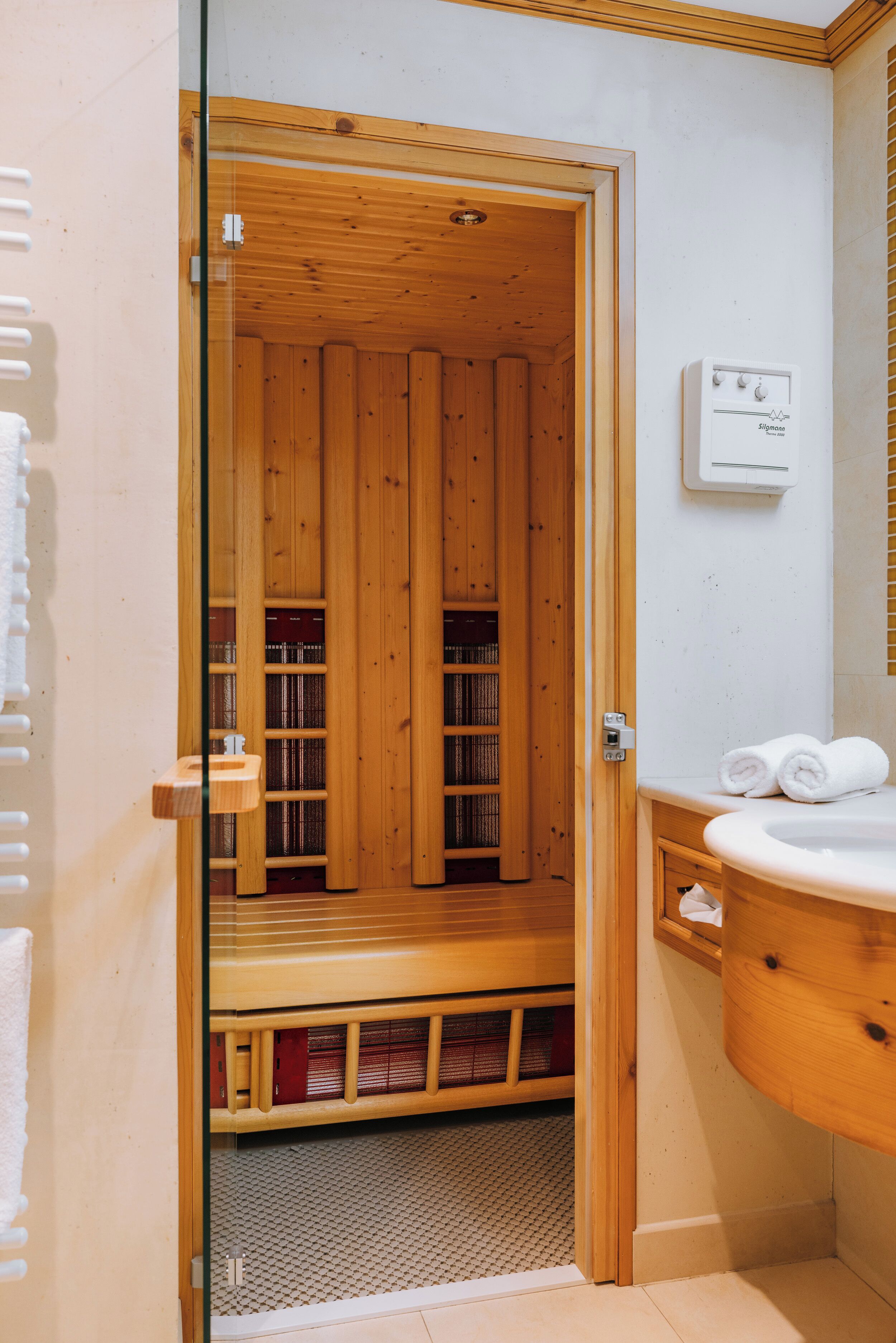 A wooden sauna room with a glass door, a white wall-mounted heater, and a wooden bench.