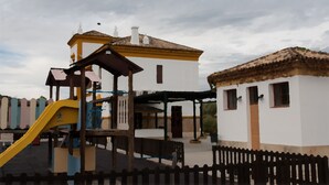 A playground with a yellow slide and a small shelter, adjacent to a white building with a brown roof and a fence. - Casa Vagón Vía Verde Sierra (Olvera)