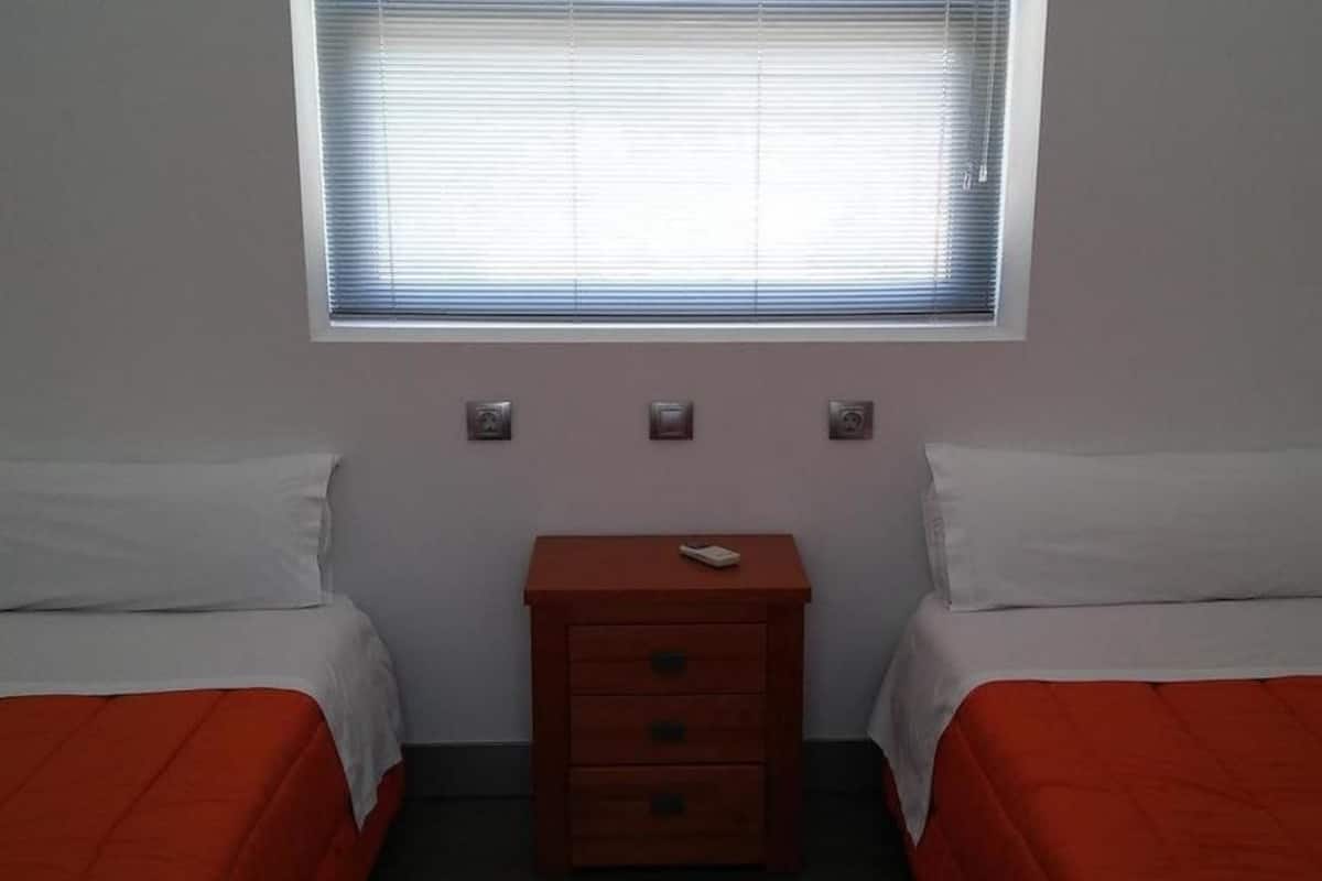 Una habitación pequeña y sencilla con dos camas individuales, una mesita de noche de madera y una ventana con persianas.