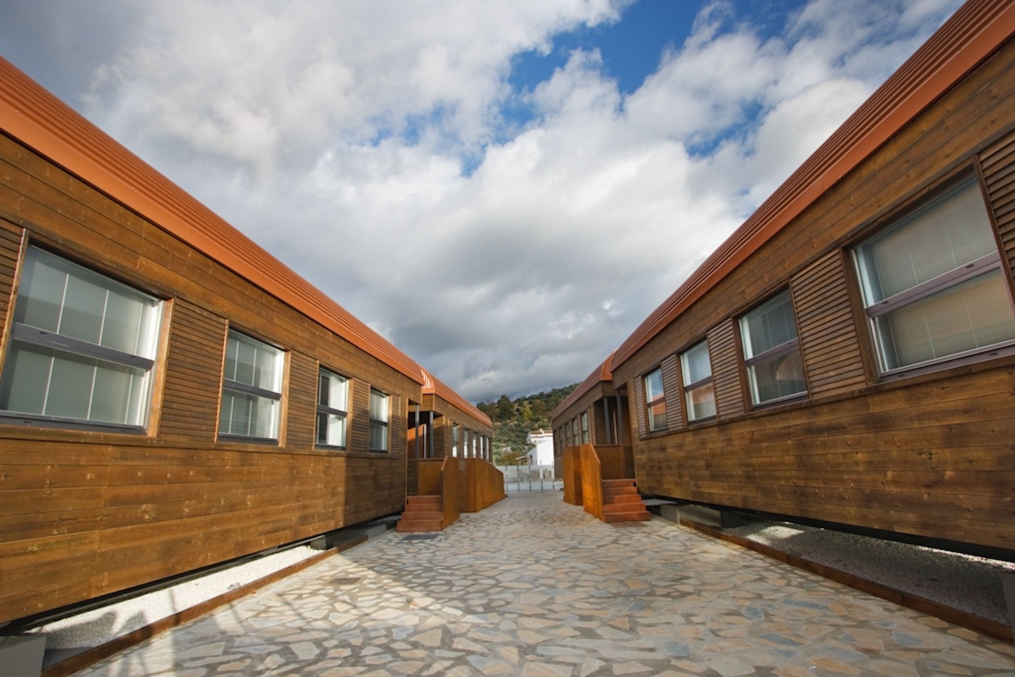 A corridor with wooden walls and stone flooring, leading to a white building in the distance.