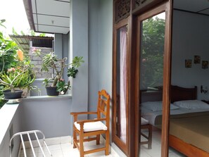 A balcony with potted plants, a wooden chair, and a sliding glass door leading to a bedroom with a bed and a nightstand.