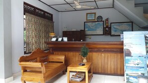A reception area with a wooden reception desk, a wooden sofa, a small table with a plant, and a wall-mounted clock. - Celagi Inn (Padangbai)
