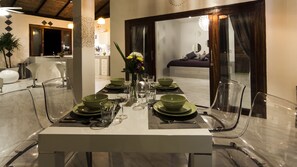 A dining table set with green plates, glasses, and cutlery, surrounded by clear chairs, in a modern interior with a view of a bed and a bathroom in the background.