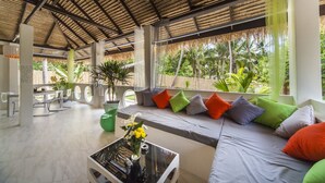 Un espacio interior amplio con techo alto, una zona de estar con cojines, una mesa de cristal con un florero, y vistas a palmeras afuera.