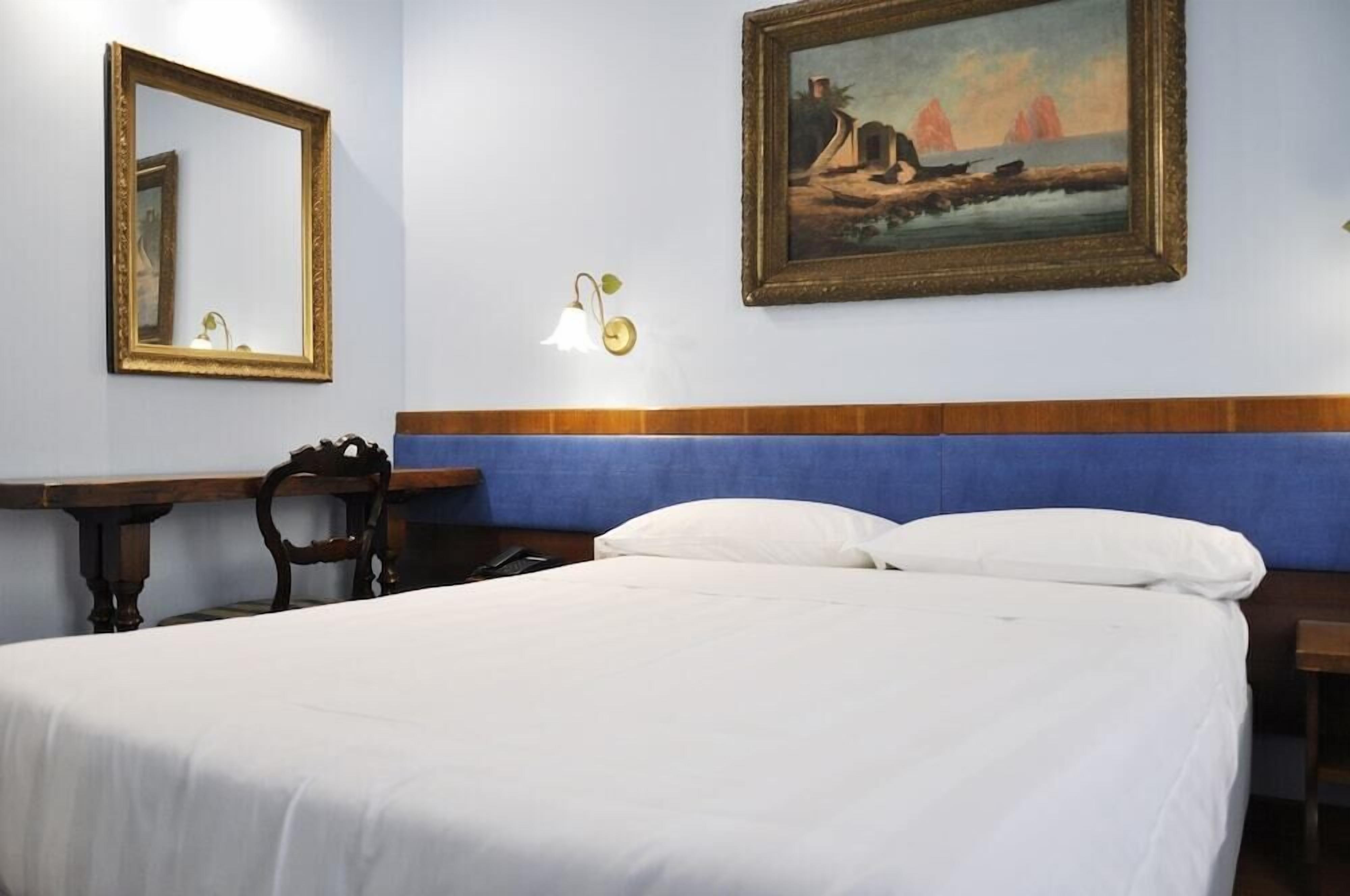 A hotel room with a bed, a wooden nightstand, a desk, a chair, a wall-mounted lamp, and a framed painting.